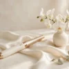 Vibromasseur baguette magique ivoire et doré posé sur soie crème près d'orchidées blanches